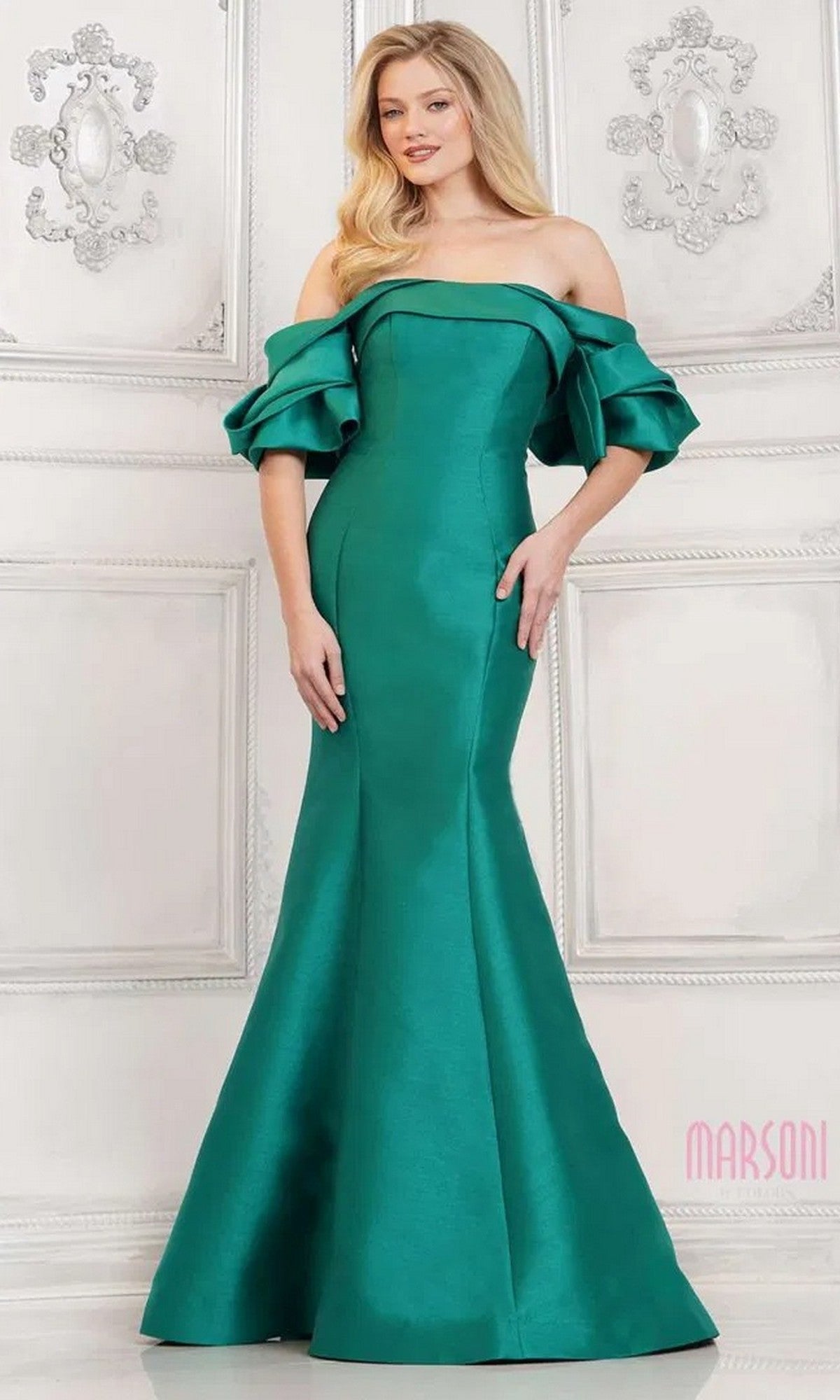 A woman wearing the Marsoni MV1290 long formal dress in emerald, featuring off-the-shoulder ruffled sleeves and a subtle mermaid silhouette, stands against an ornate white wall.